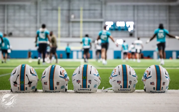 Miami Dolphins players warm up in the background, in the forefront are five player helmets
