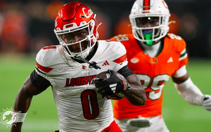 Louisville wide receiver in a white jersey, No. 0, sprints downfield carrying the football while being pursued by a Miami Hurricanes defender in an orange jersey, No. 89, during a college football game.